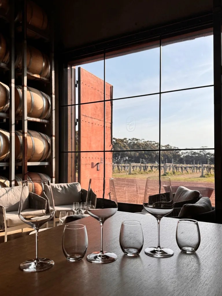 View of wine glasses arranged on a table inside a winery, with wooden barrels in the background and a scenic vineyard visible through large windows.