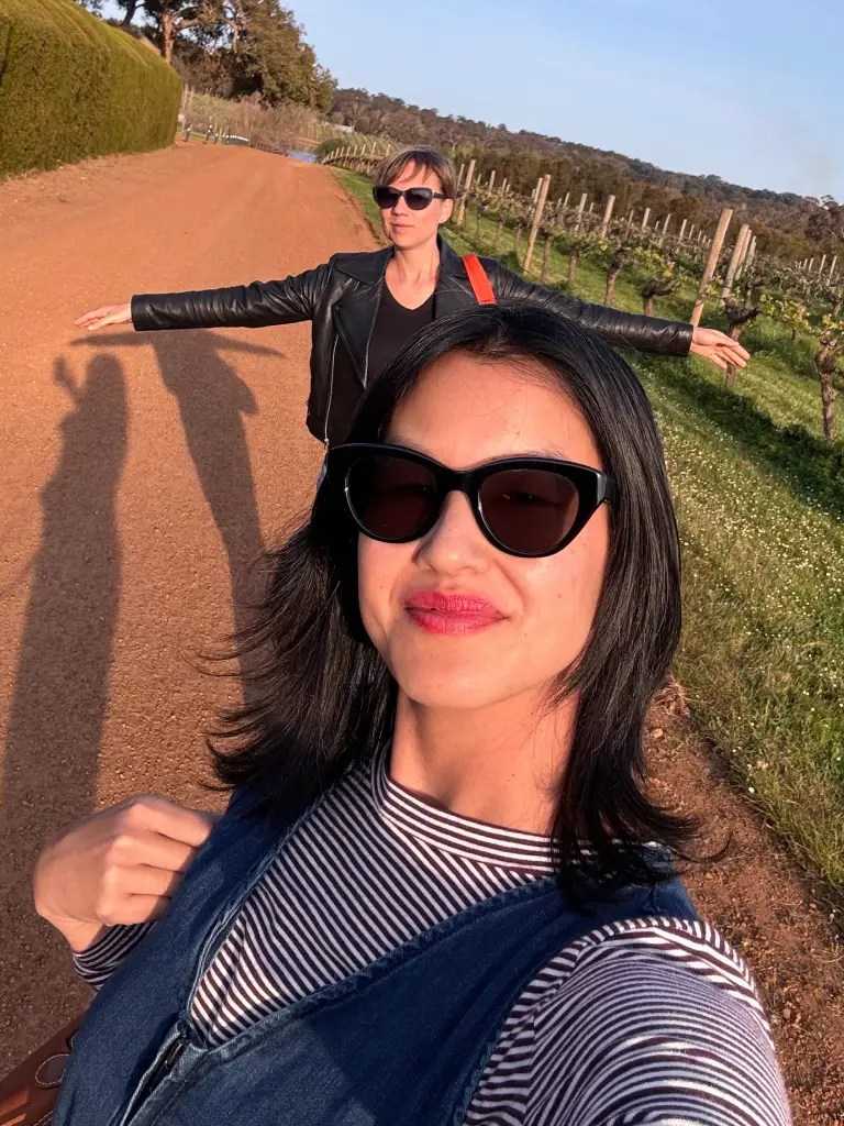 Two women enjoying a vineyard walk, posing for a selfie on a dirt path lined with grapevines and greenery in the background.