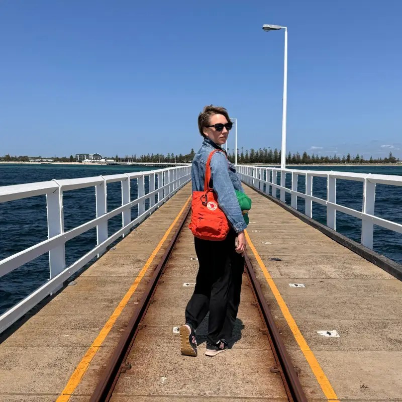A woman stands on Busselton Jetty, looking back toward the camera, wearing sunglasses and a denim jacket, with a red bag slung over their shoulder. The background features a clear blue sky and the ocean.