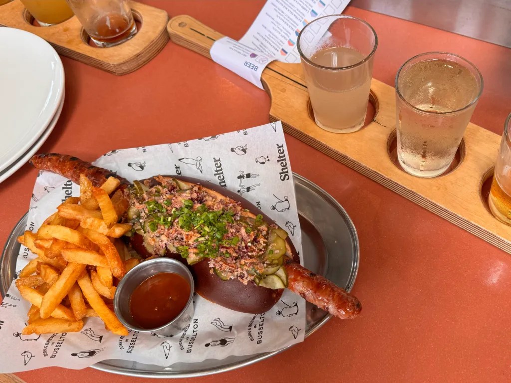 A plate of food featuring a sausage in a bun topped with herbs and pickles, accompanied by fries and a small bowl of sauce, with a flight of drinks in the background.