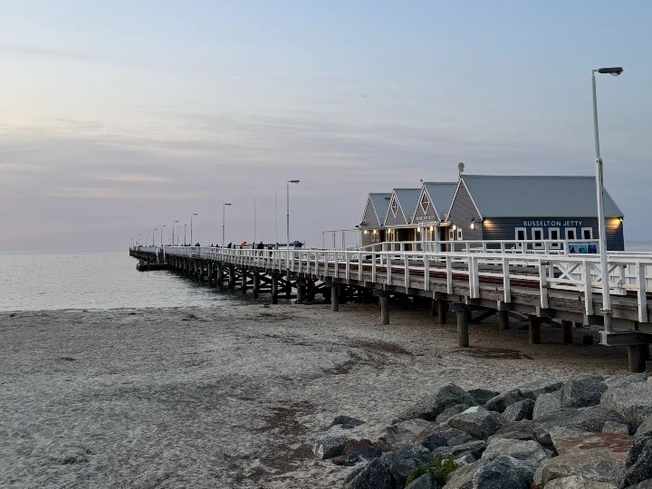 View of Busselton Jetty at sunset, showcasing the long wooden pier extending into the calm waters, with a building at the end and sandy beach in the foreground.