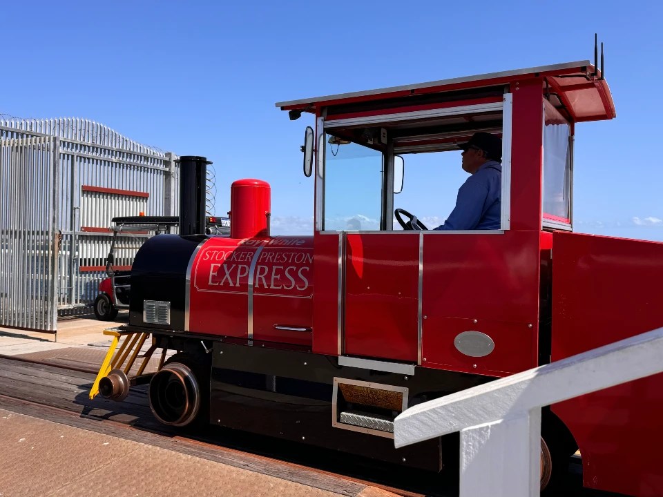 A small red train named 'Stocker Preston Express' is parked on a platform, with a conductor visible inside the cab, set against a clear blue sky and surrounded by a fence.