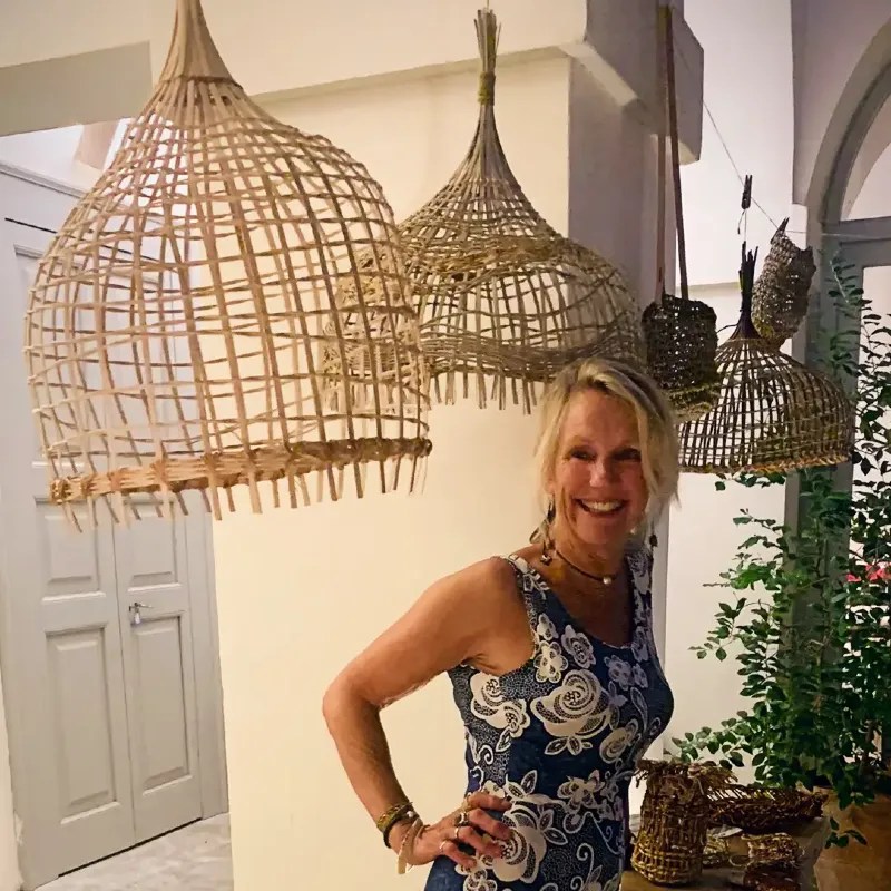 A smiling woman posing next to woven baskets hanging on a wall, showcasing her artistry in a cozy interior space.