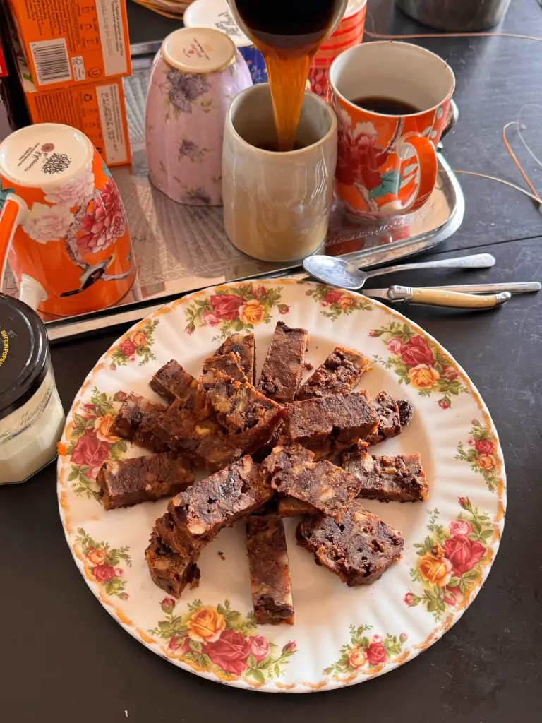 A plate with slices of dessert bars made from nuts and chocolate, arranged in a circular pattern, with cups and a coffee pot in the background.