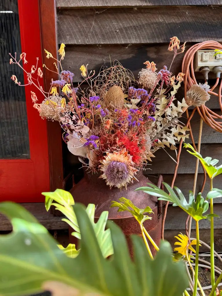 A vintage pot filled with an arrangement of dried flowers and foliage, with vibrant colors and textures, set against a rustic wooden wall.