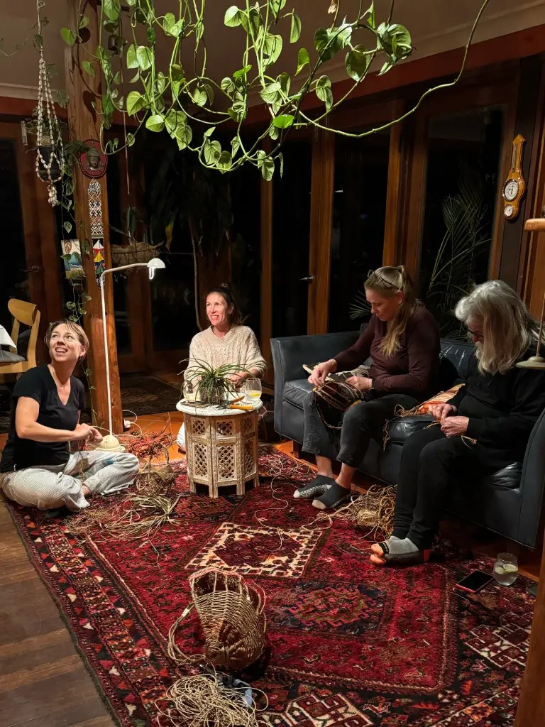 A cozy indoor setting with four women engaged in weaving activities, surrounded by natural materials and plants hanging from the ceiling. The room features a warm atmosphere with a patterned rug and a small table at the center.