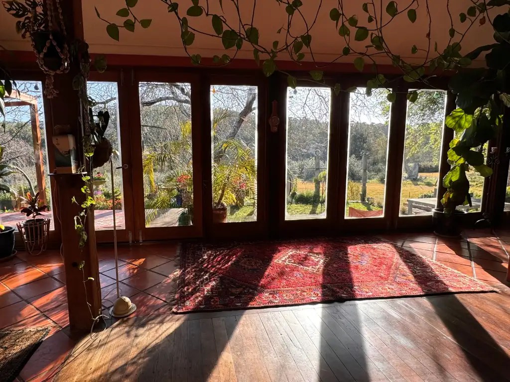 Interior view of a cozy room with large windows allowing natural light, showcasing a rustic charm with wooden flooring and a patterned rug. The background features lush greenery and outdoor plants visible through the windows.