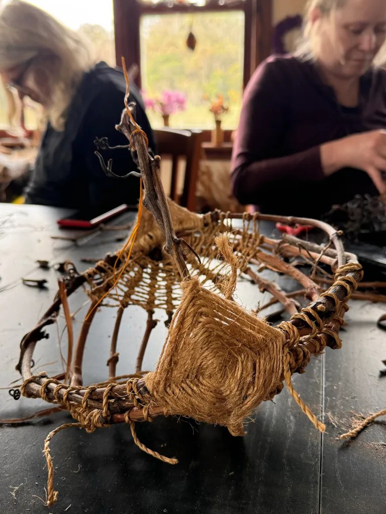 A handmade woven melon basket made from natural fibers, showcased on a table during a crafting workshop, with two women in the background engaged in their own craft activities.