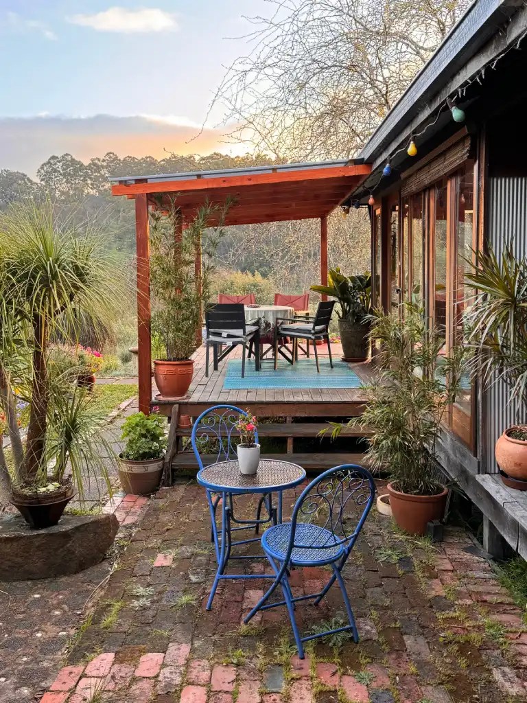 A cozy outdoor seating area with two blue bistro chairs and a small table, surrounded by plants and potted flowers. The wooden deck leads to a rustic house with large windows, and the background shows a picturesque landscape under a soft sunset.