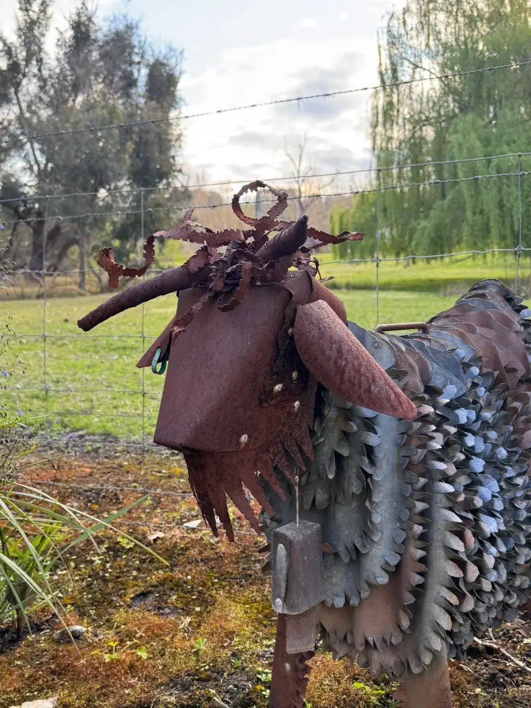 A whimsical metal sculpture resembling a goat, with various textured metal elements for its body and head, set against a green field and trees in the background.
