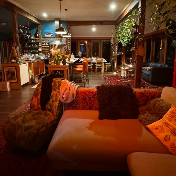 Cozy interior of a rustic living space with a kitchen in the background, featuring a large sofa adorned with colorful pillows and throws, ambient lighting, and a dining area set for a meal.