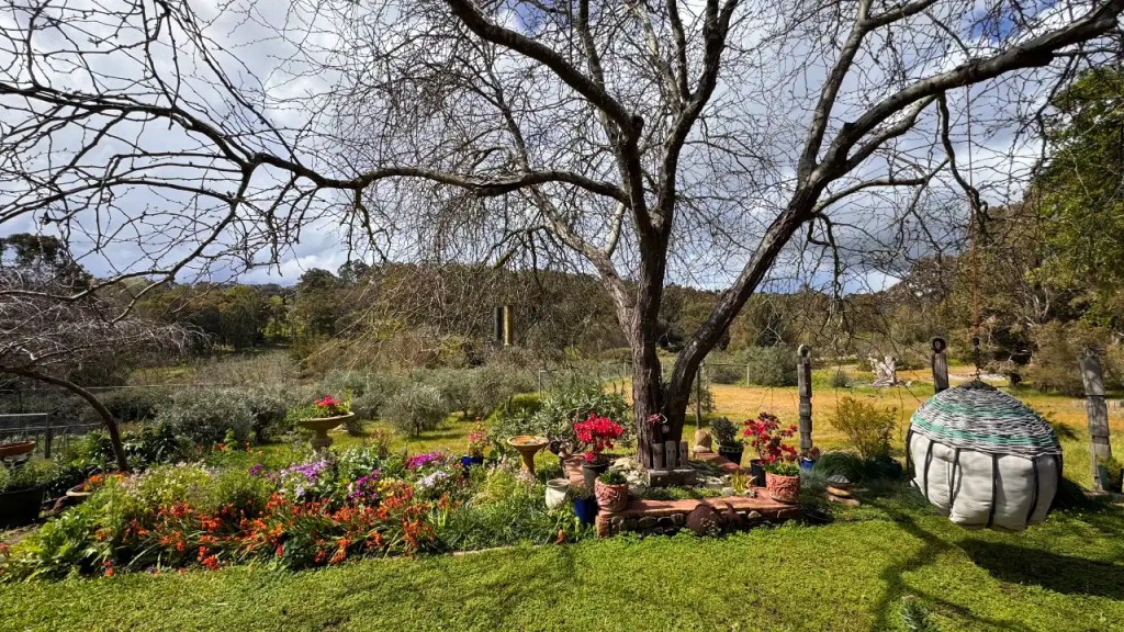 A serene garden scene at Kangamia retreat in Balingup, featuring colorful flowers, a decorative birdbath, and a large tree with bare branches, under a cloudy sky.
