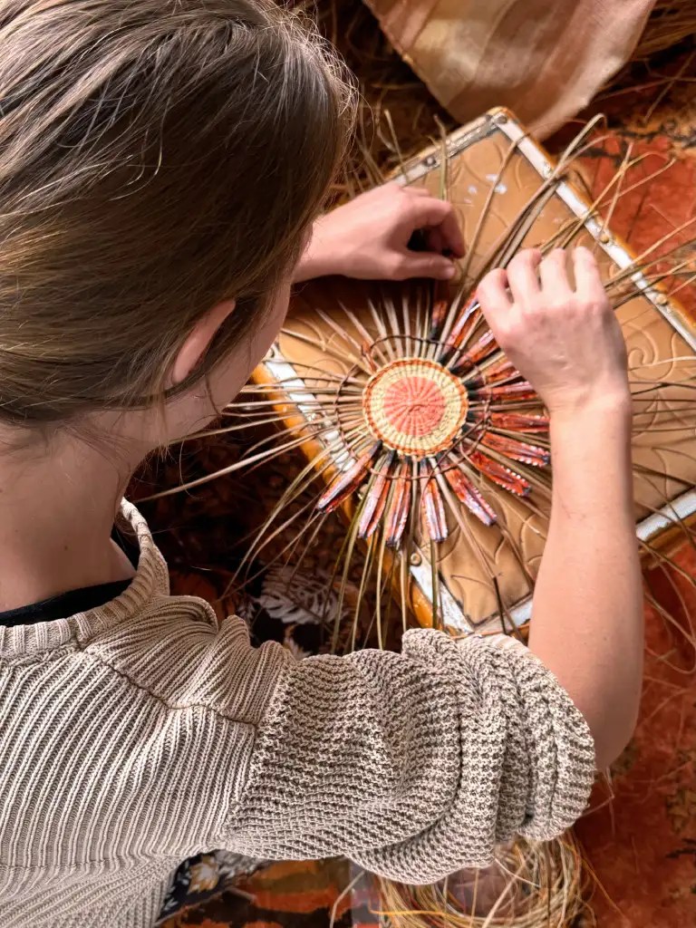 A person working on a woven basket, using natural materials and focused on the intricate design.