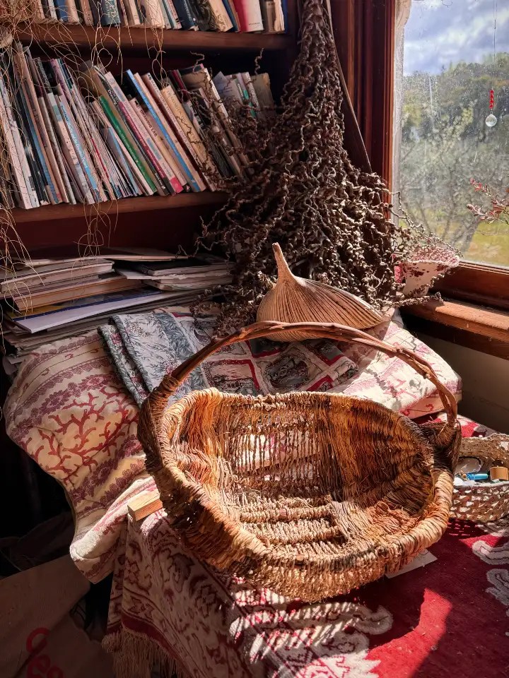 A handmade woven basket on a decorative table next to a stack of books, with sunlight streaming through a window.