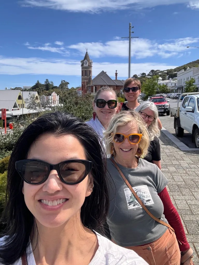 A group of five women taking a selfie outdoors with a historic building in the background, enjoying a sunny day.