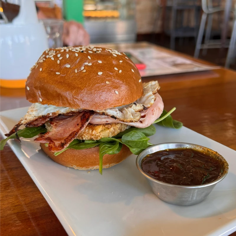 A gourmet breakfast roll featuring a sesame seed bun, filled with leafy greens, crispy bacon, a fried egg, and ham, accompanied by a small dish of tomato chutney on the side.