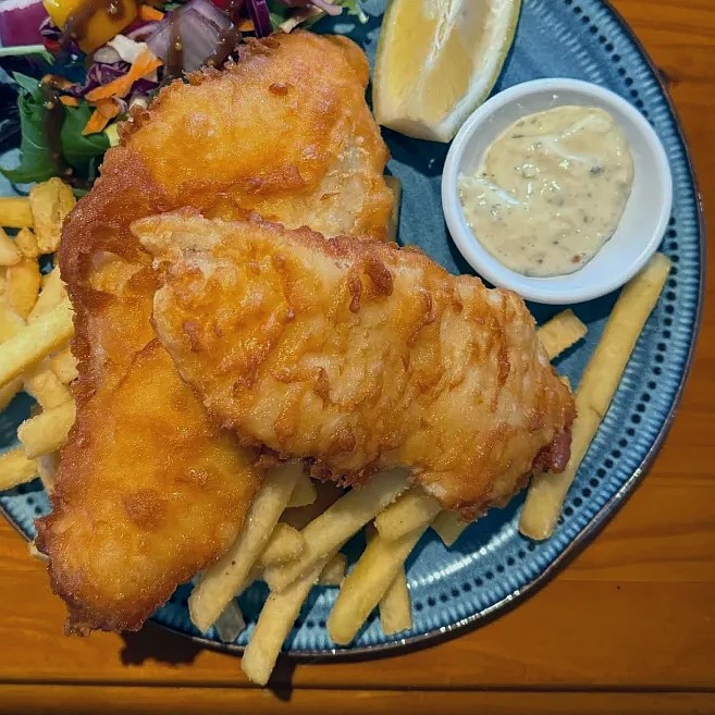 A plate featuring two pieces of golden-battered fish fillets served with French fries, a small bowl of dipping sauce, a lemon wedge, and a side salad with colorful shredded vegetables.