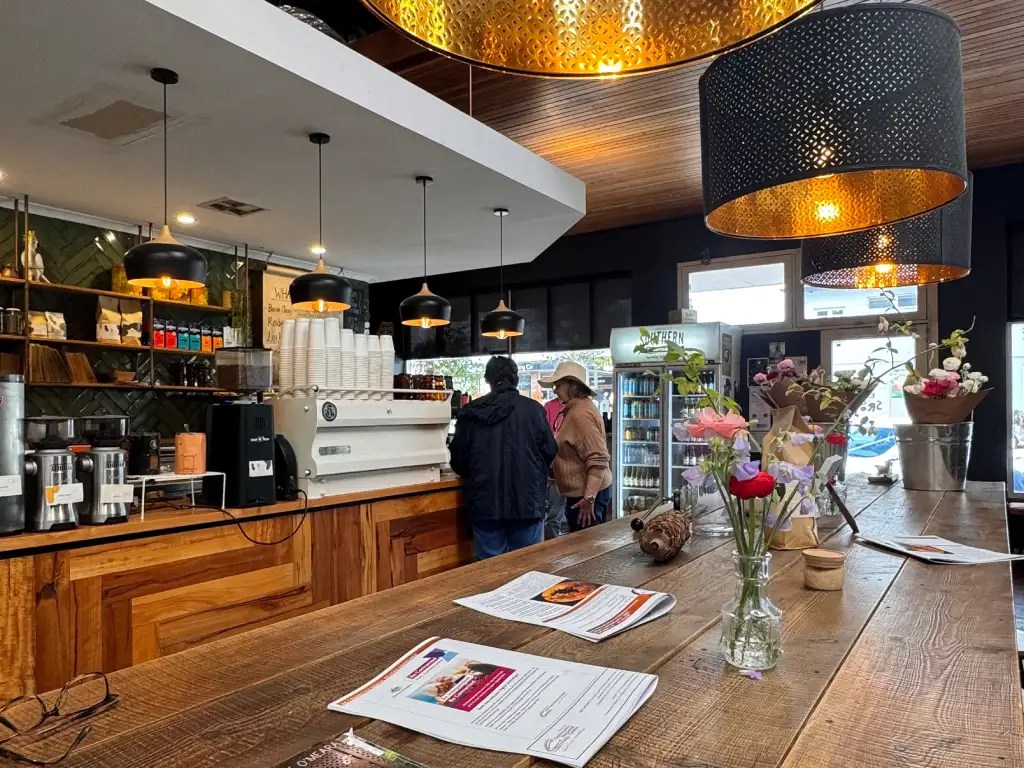Interior of a cozy café featuring a wooden counter, coffee equipment, and decorative hanging lights. Patrons are interacting at the counter, while flowers in vases add a touch of charm.