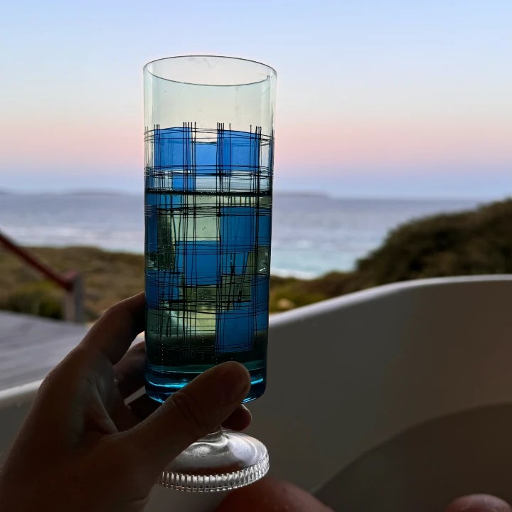 A hand holding a blue and clear patterned glass filled with prosecco in a spa tub, set against a backdrop of a coastal sunset with soft pink and blue hues in the sky.