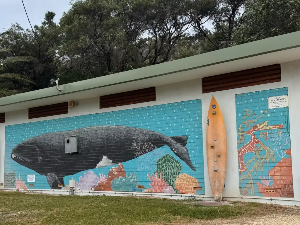 A colorful mural depicting a whale surrounded by marine life and coral, painted on a building wall, with a surfboard leaning against it.