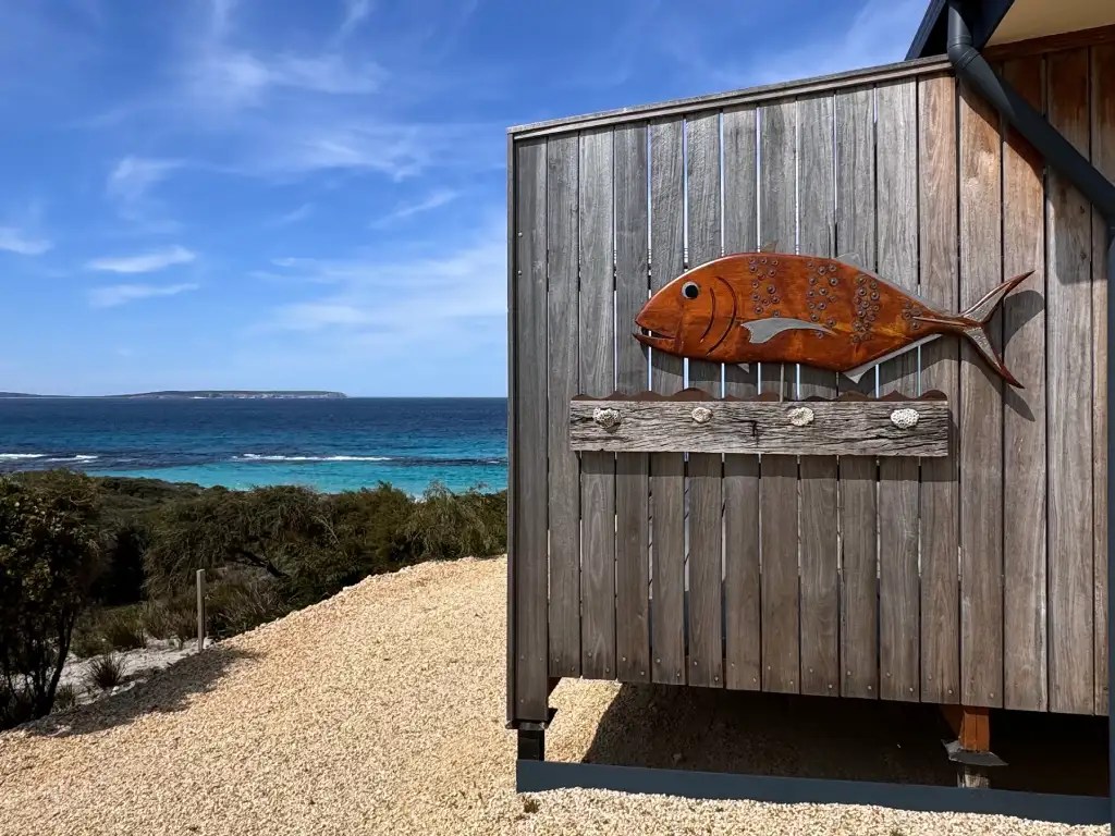 A wooden wall featuring a fish sculpture, overlooking a beautiful coastal view with turquoise waters and a clear blue sky in Bremer Bay, Western Australia.