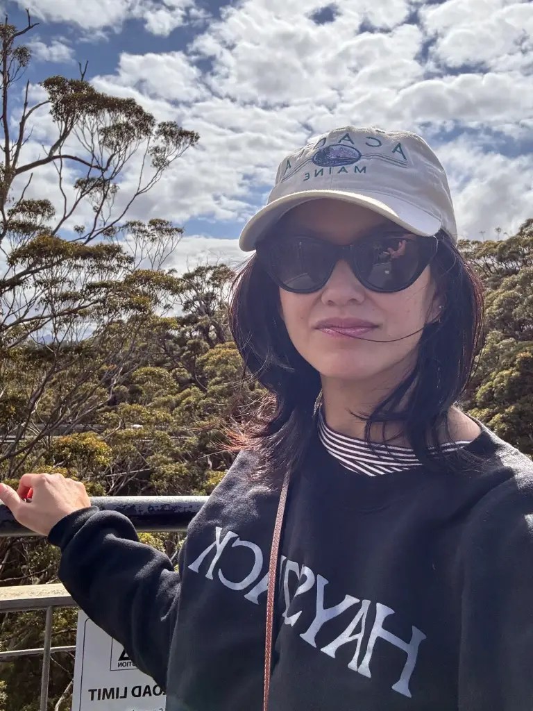 A person wearing sunglasses and a cap stands on a railing, posing for the camera with trees and a cloudy sky in the background.