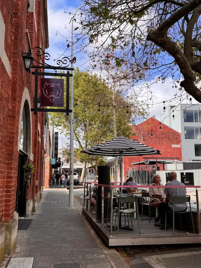 Street view of a brick building with a storefront sign featuring a tiger logo. Outdoor seating is visible where a couple sits at a table. The scene is framed by trees with few leaves, and a cloudy sky above.
