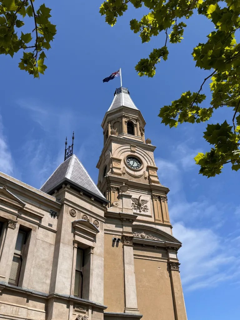 Fremantle Town Hall's historic clock tower with a flag flying, surrounded by green leaves against a bright blue sky.