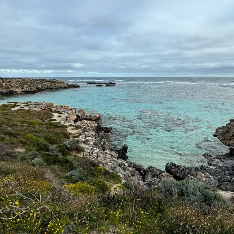 Scenic view of Rottnest Island's coastline featuring turquoise waters, rocky shores, and patches of greenery under a cloudy sky.