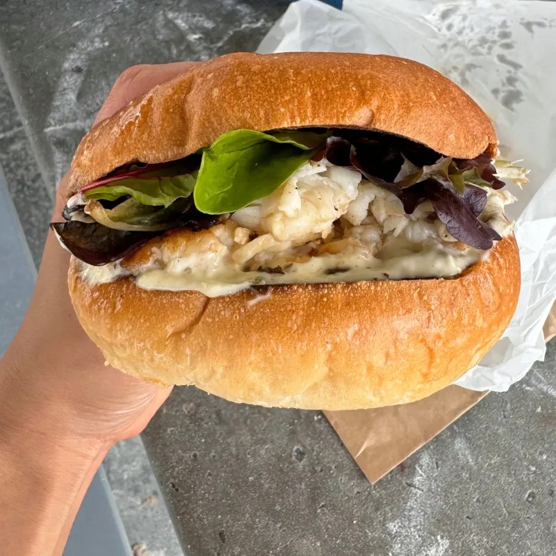 A close-up of a crayfish roll sandwich with fresh greens, held by a hand. The sandwich features soft bread, filled with grilled crayfish and creamy sauce, surrounded by leafy greens.