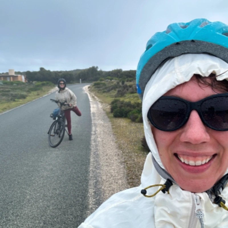 Two cyclists enjoying a rainy day on Rottnest Island, smiling for a selfie while one poses next to their bike on a quiet road surrounded by nature.