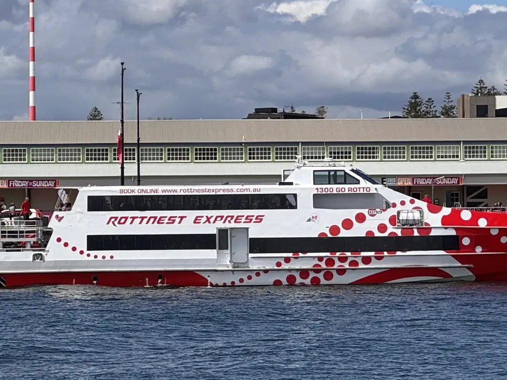 A high-speed ferry named 'Rottnest Express' docked at a terminal, with red and white designs visible on its side, under a partly cloudy sky.