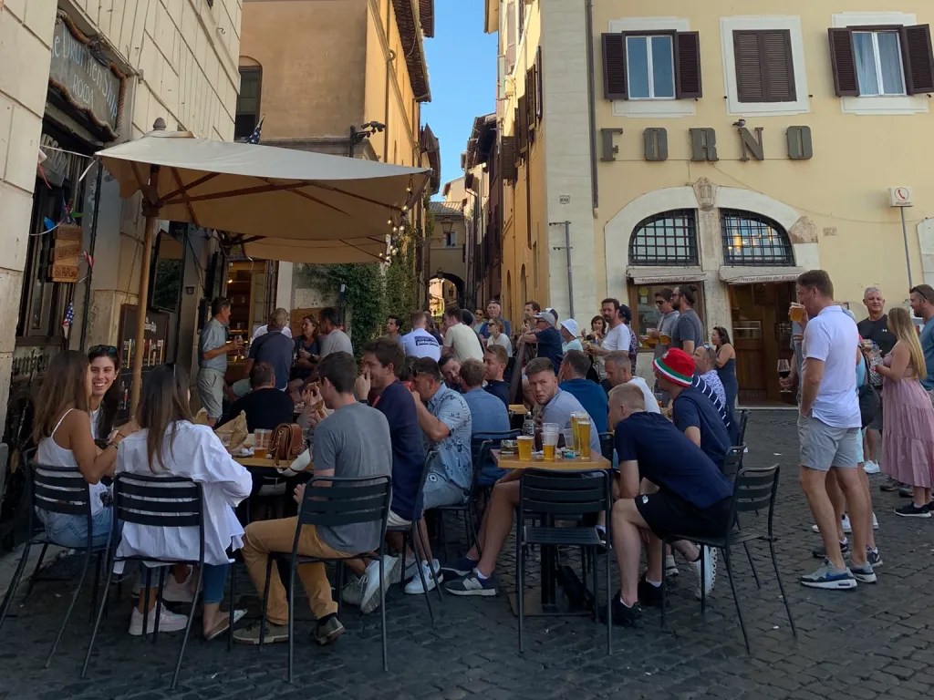 A bustling outdoor scene at Campo de Fiori in Rome, with people gathered around tables enjoying drinks and food on a sunny day.