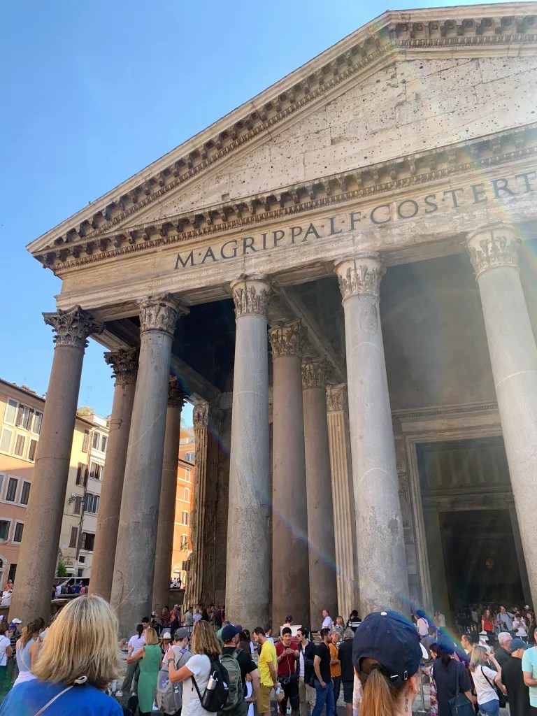 The Pantheon in Rome, featuring its iconic columns and a crowd of visitors gathered under a clear blue sky.