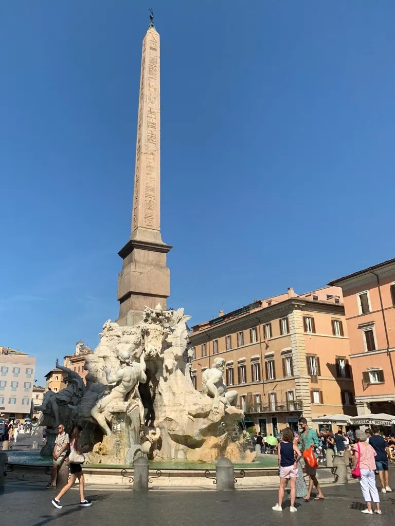 Piazza Navona featuring the Fountain of the Four Rivers and an ancient obelisk, surrounded by visitors and historic buildings under a clear blue sky.