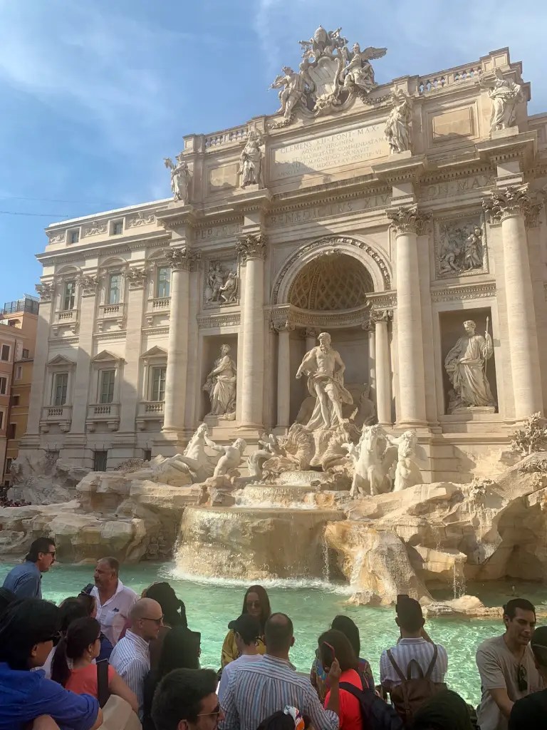 Crowd gathering around the Trevi Fountain in Rome, showcasing the ornate Baroque architecture and intricate sculptures of the fountain.