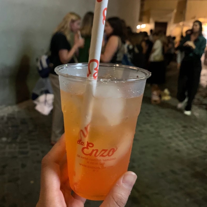 A person holding a plastic cup with an Aperol Spritz and ice, featuring a straw with the logo of a restaurant called Da Enzo, while a group of people is blurred in the background.