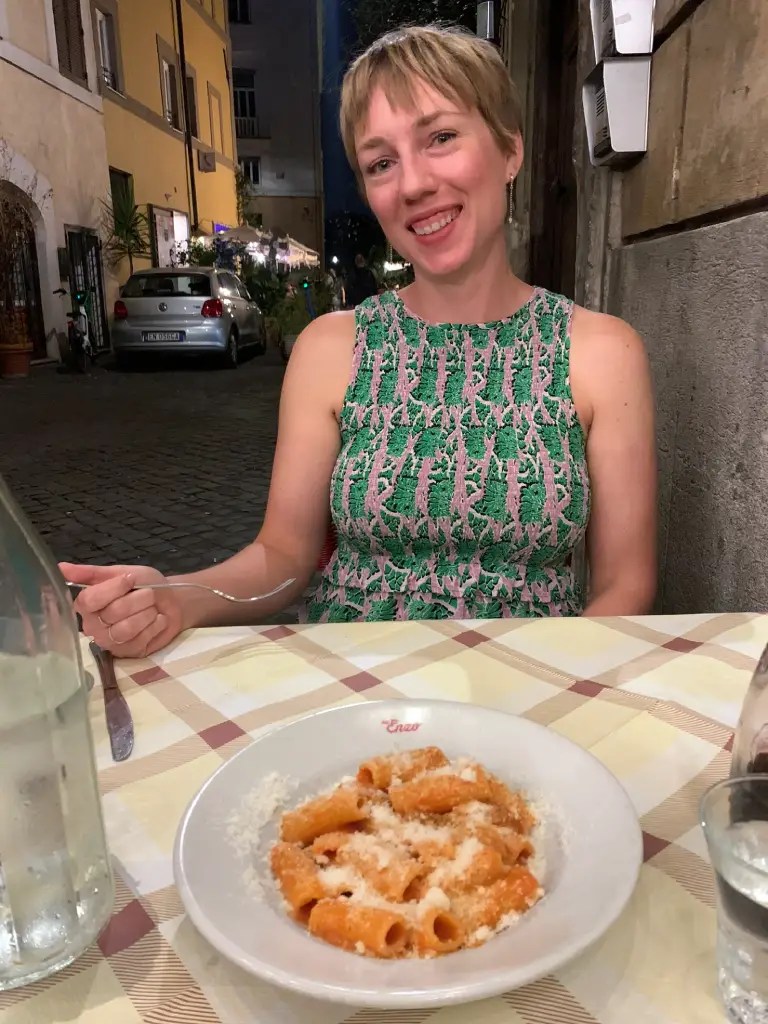 Person sitting at a table in a restaurant, smiling and holding a fork, with a plate of pasta in front of them. A bottle of water and a glass are also visible on the table.