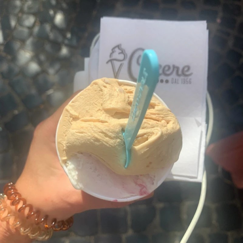 A close-up of a hand holding a cup of gelato featuring a creamy scoop of caramel-colored gelato atop a white scoop with a red swirl, with a blue plastic spoon. A paper napkin with a logo is visible in the background, and cobblestones can be seen beneath.