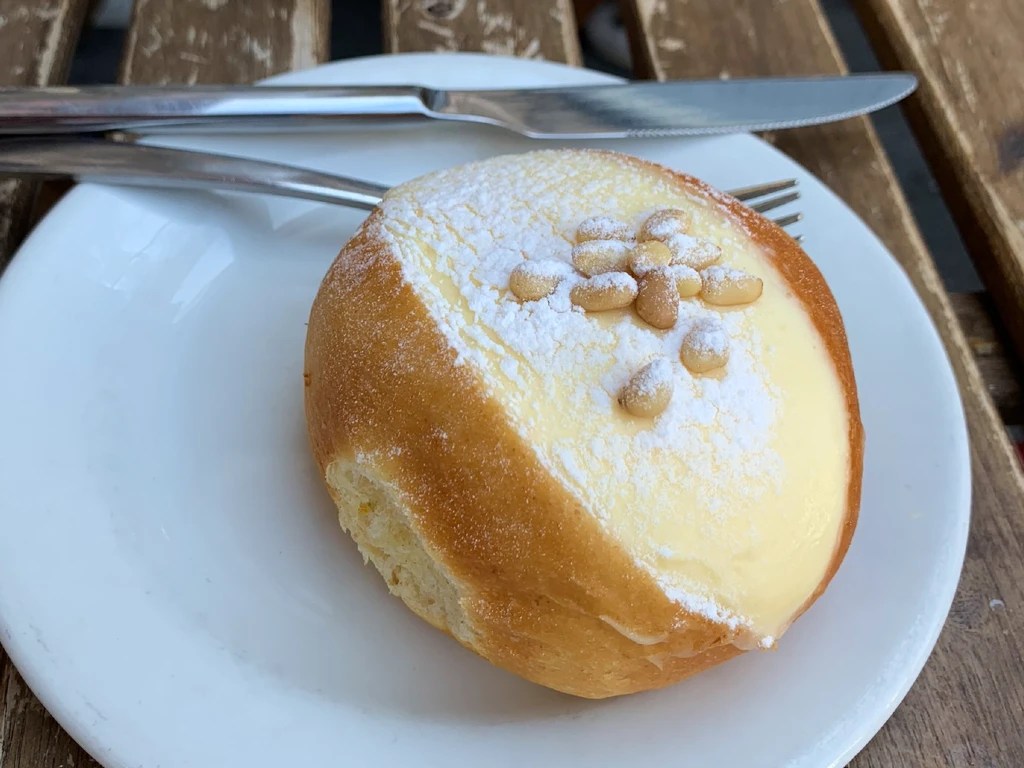 A plate with a sweet Italian bun filled with cream and topped with pine nuts, alongside a knife and fork on a wooden table.