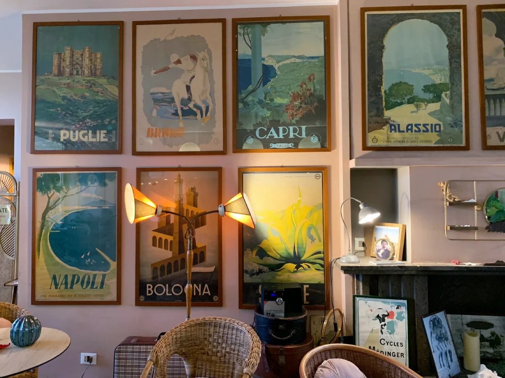 A cozy interior featuring vintage travel posters from various Italian destinations, including Puglia, Napoli, Capri, and Bologna, alongside a stylish lamp and wicker seating.