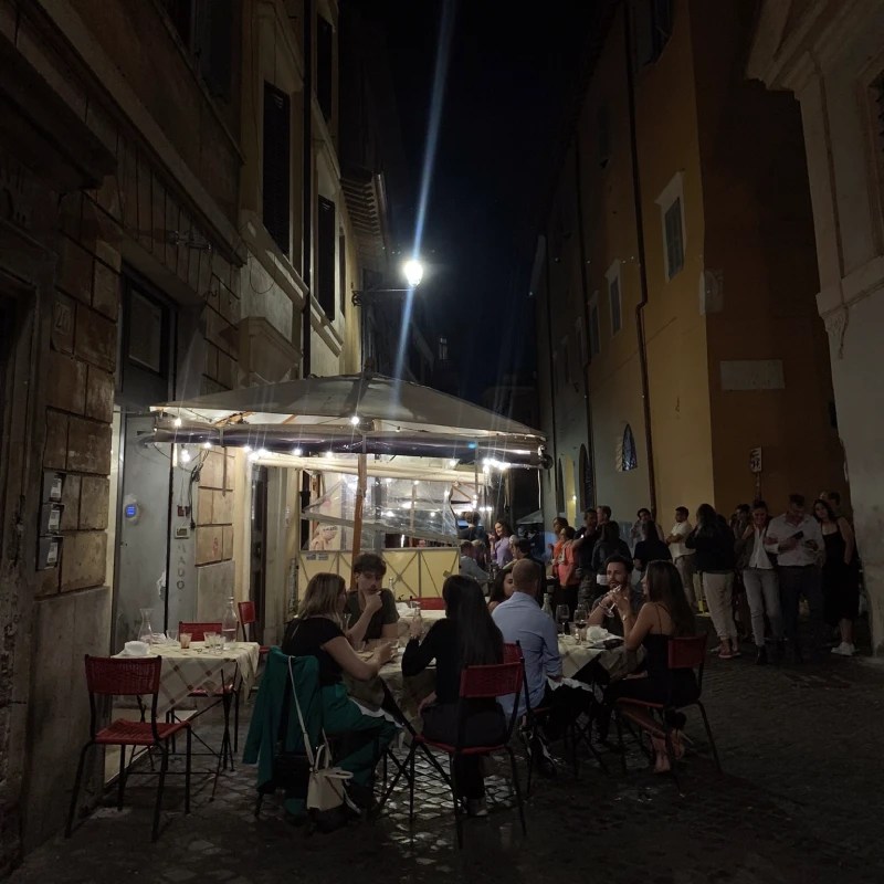 An outdoor dining scene at night in a narrow Roman street, featuring tables set with diners enjoying their meals, illuminated by string lights and street lamps.