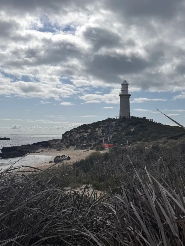 View of Bathurst Lighthouse in Rottnest Island, perched on a hill under a cloudy yet clearing sky, with thick wild grasses in the foreground.