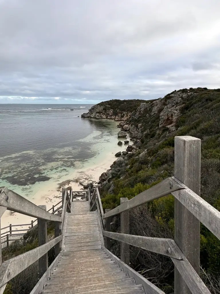 A wooden staircase leading down to Porpoise Bay on Rottnest Island, surrounded by lush greenery and rocky coastline under a cloudy sky.
