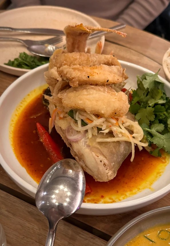 A plate of fried whole fish topped with crispy fried squid and served with a side of fresh herbs, chili, and a tangy sauce.